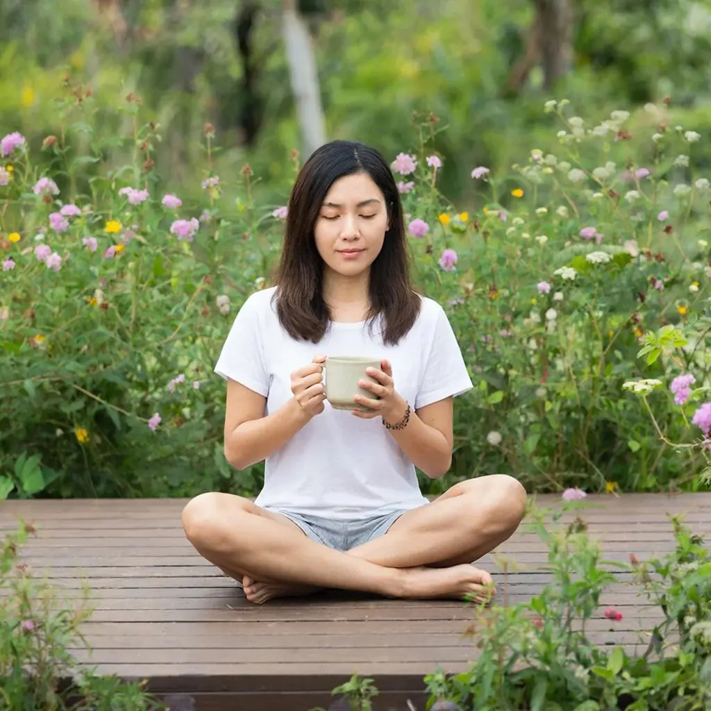A woman of Asian descent sitting peacefully in a garden, holding a ceramic mug, representing natural hormonal balance.